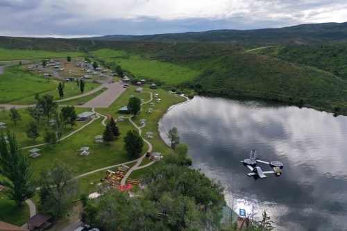 Aerial view of a serene lake surrounded by green hills, with campsites and kayaks along the shore.