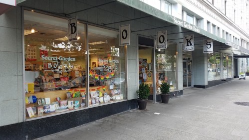 A bookstore named "Secret Garden Books" with large windows displaying colorful books and a welcoming entrance.