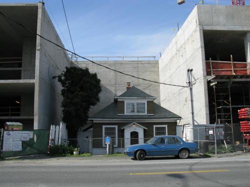 A small house stands between two large concrete buildings under construction, with a blue car parked in front.
