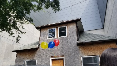 A small, weathered house with a slanted roof, surrounded by modern buildings, holding colorful balloons.