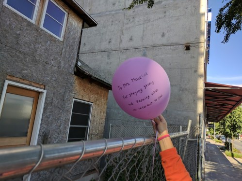 A hand holds a pink balloon with a message, near a construction site and a fence.