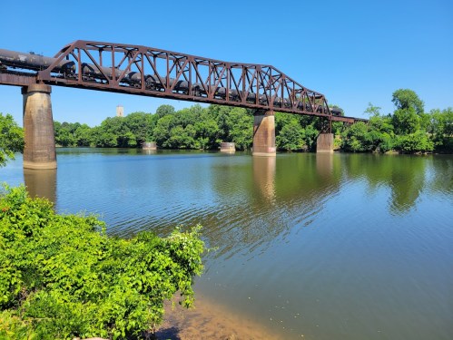 A train bridge spans a calm river, surrounded by lush greenery and a clear blue sky. Reflections shimmer on the water.