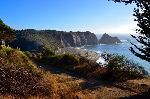 Scenic coastal view with cliffs, a beach, and ocean waves under a clear blue sky. Lush greenery in the foreground.