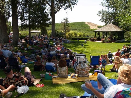 A large crowd sits on the grass at an outdoor event, enjoying the scenery and a gazebo in the background.