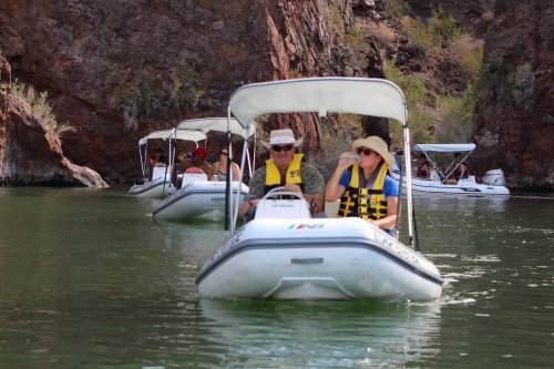 Two people in a small boat wearing life jackets, navigating a calm green lake surrounded by rocky cliffs.