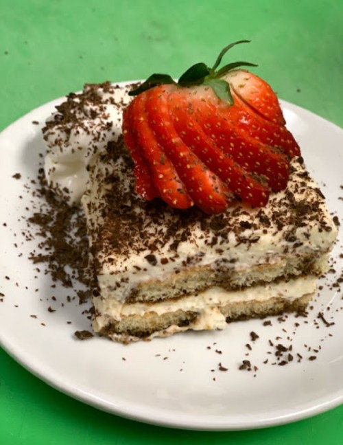 A slice of dessert topped with a strawberry, chocolate shavings, and whipped cream on a green background.