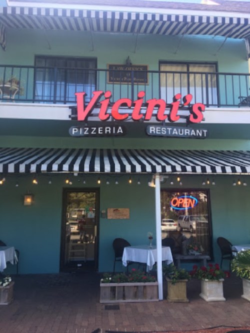 Exterior of Vicini's Pizzeria, featuring a blue facade, striped awning, and an "OPEN" sign. Tables are set outside.