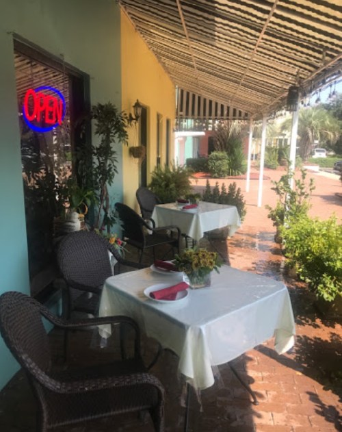 Outdoor dining area with tables set for guests, a bright "OPEN" sign, and lush greenery in a sunny setting.