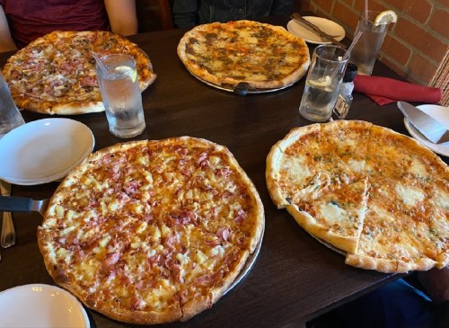 Four pizzas on a table, with various toppings, alongside glasses of water and empty plates.
