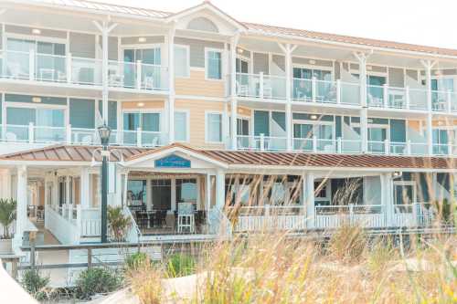 A coastal hotel with balconies, surrounded by tall grass and a clear sky, featuring a welcoming entrance.