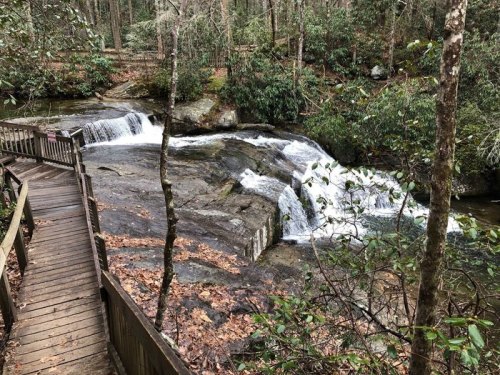 A wooden walkway beside a serene waterfall surrounded by lush greenery and rocky terrain.