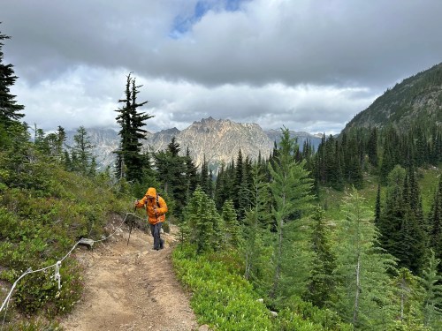 A hiker in an orange jacket walks along a trail surrounded by lush greenery and mountains under a cloudy sky.