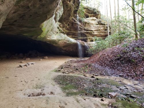 A serene cave with a small waterfall, surrounded by lush greenery and sandy ground.