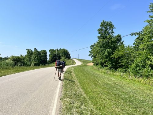 A person walks along a rural road, carrying a backpack, with green trees and a clear blue sky in the background.