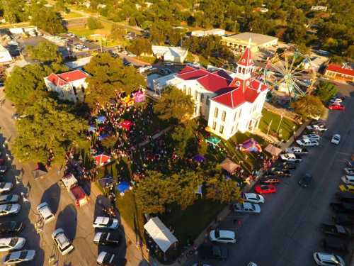 Aerial view of a festive gathering with a historic building, carnival rides, and crowds in a park-like setting.