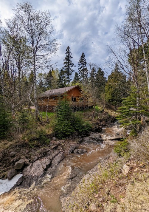 A wooden cabin nestled among trees beside a flowing creek, with a cloudy sky overhead.