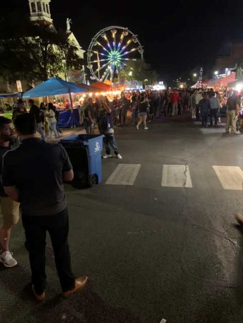 A lively night market scene with crowds, food stalls, and a colorful Ferris wheel in the background.