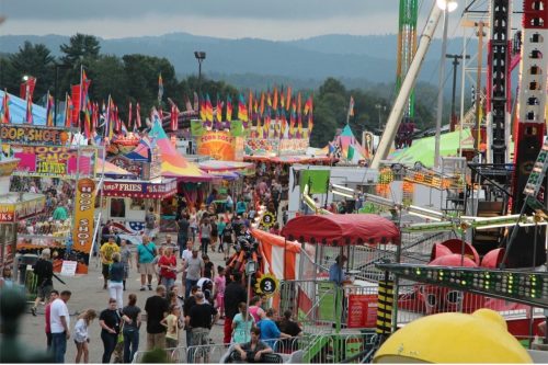 A vibrant carnival scene with colorful tents, flags, and crowds enjoying rides and games under a cloudy sky.