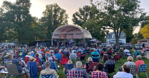 A large crowd sits on lawn chairs at an outdoor concert under a canopy, surrounded by trees and parked cars.