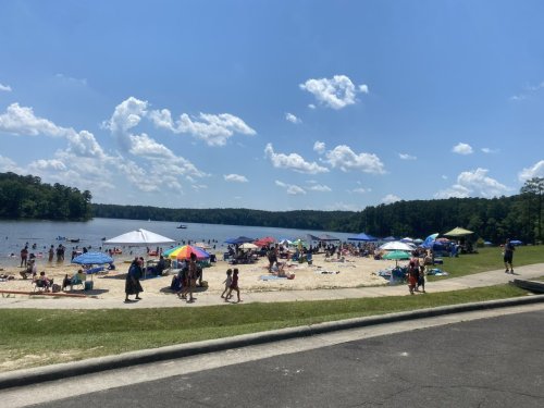 A busy beach scene with people enjoying the sun, umbrellas, and a lake under a bright blue sky with fluffy clouds.