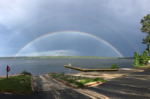 A double rainbow arcs over a calm lake, with dark clouds in the background and a wooden dock in the foreground.