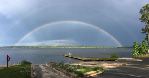 A vibrant double rainbow arcs over a calm lake, with a wooden dock and trees in the foreground.