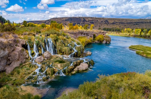 A scenic view of cascading waterfalls surrounded by lush greenery and a calm river under a blue sky with clouds.