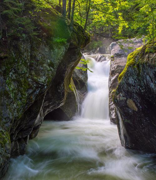A serene waterfall cascades between moss-covered rocks, surrounded by lush green trees in a tranquil forest setting.