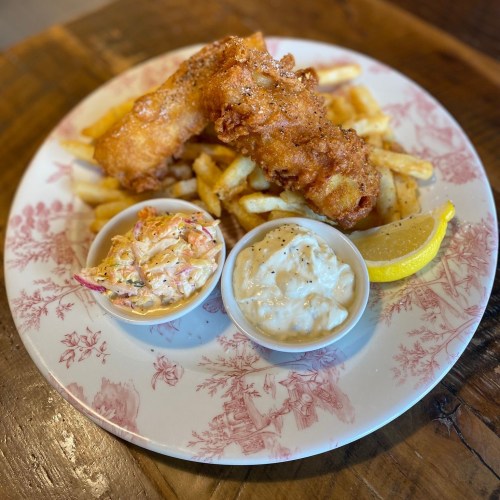 A plate with crispy fish fillets, golden fries, coleslaw, tartar sauce, and a lemon wedge on a decorative dish.