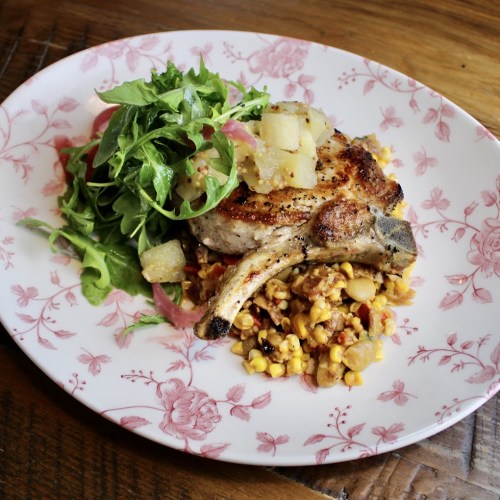 A grilled pork chop topped with fruit, served with a side of arugula salad and a colorful grain medley on a floral plate.
