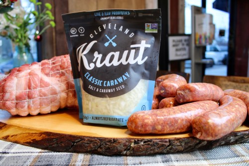 A wooden board displays a bag of Cleveland Kraut, sausages, and a piece of meat, set in a rustic kitchen.