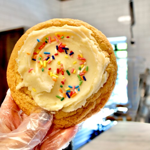 A hand holding a large cookie topped with creamy frosting and colorful sprinkles.