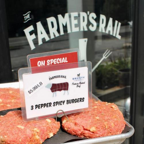 A sign displays "3 Pepper Spicy Burgers" for $5.90/lb outside The Farmer's Rail, with burger patties in the foreground.