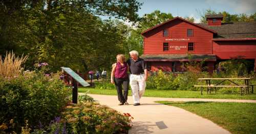 A couple walks hand in hand along a path in a park, with a red barn and flowers in the background.