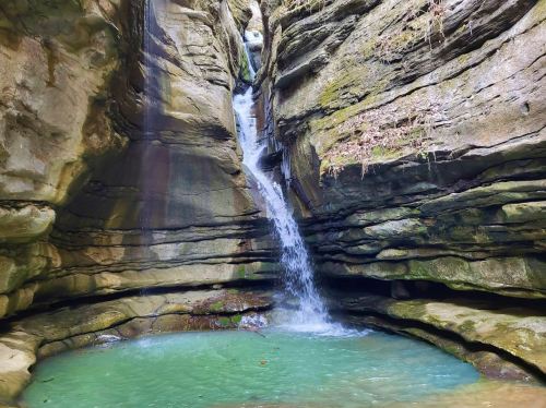 A serene waterfall cascades into a clear pool, surrounded by rugged stone walls and lush greenery.