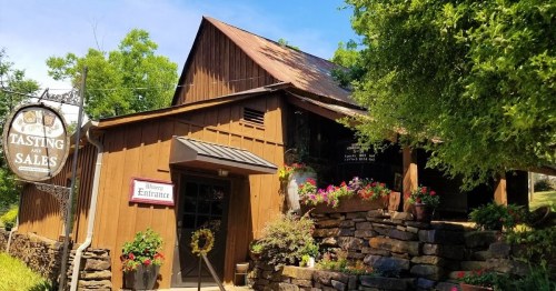 A rustic wooden building with a sign for "Tasting and Sales," surrounded by greenery and colorful flowers.
