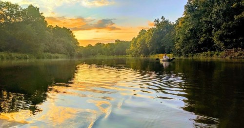 A serene river scene at sunset, with trees lining the banks and a small boat gently floating on the calm water.