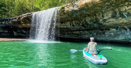 A person on a paddleboard gazes at a waterfall cascading into a turquoise lake surrounded by lush greenery.