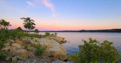 A serene lakeside view at sunset, featuring rocky shores and lush greenery under a colorful sky.