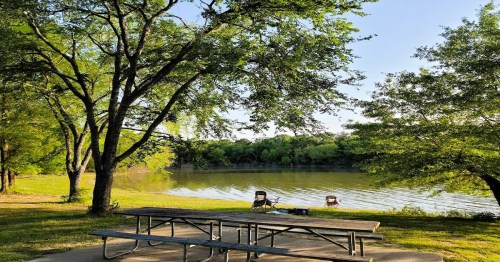 A serene lakeside scene with a picnic table and chairs under trees, reflecting calm water and greenery.