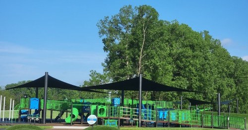 A colorful playground with green and blue equipment, shaded by large canopies, set against a backdrop of trees.