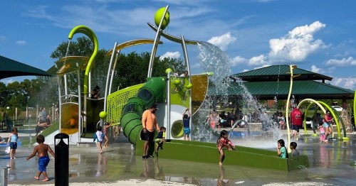 A vibrant splash pad with water features, children playing, and adults supervising under a sunny sky.