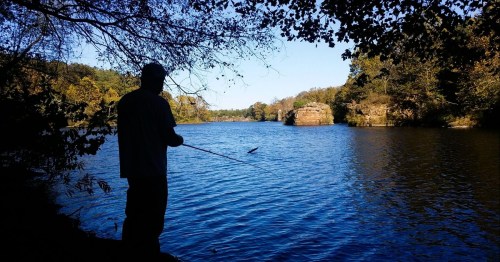 A silhouette of a person fishing by a calm river surrounded by trees and rocky formations under a clear blue sky.