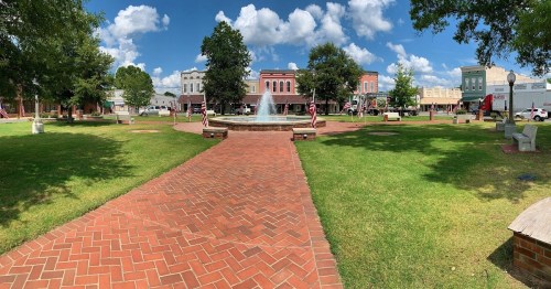 A sunny town square featuring a fountain, brick pathways, green grass, and historic buildings with American flags.