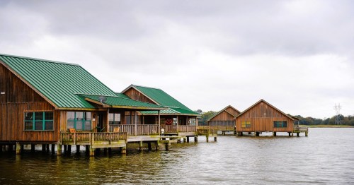 Wooden cabins with green roofs on stilts over calm water, under a cloudy sky.