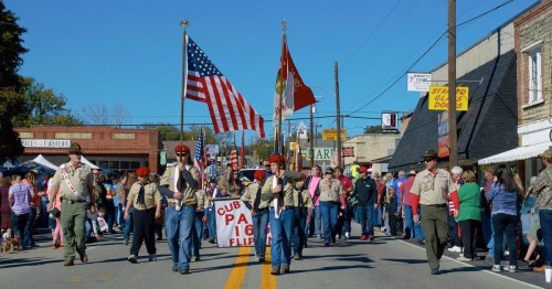 A parade with scouts carrying flags and a banner, marching down a street lined with spectators and shops.