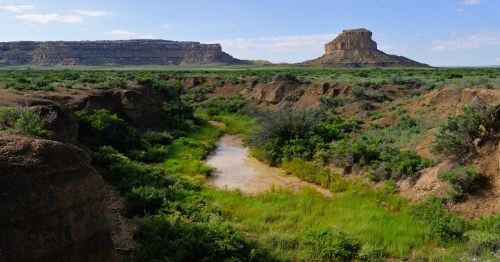 A scenic landscape featuring a winding creek surrounded by green vegetation and rocky cliffs under a blue sky.