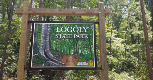 Sign for Logoly State Park surrounded by lush green trees and a dirt path leading into the forest.