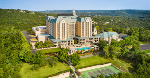 Aerial view of a large hotel surrounded by greenery, featuring a pool and tennis courts in the foreground.