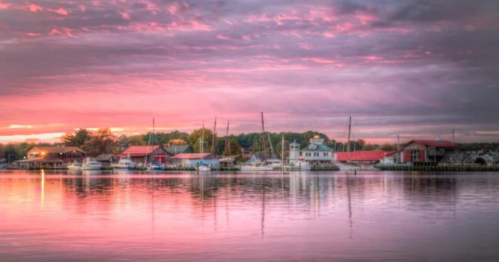 A serene harbor at sunset, with colorful skies reflecting on calm waters and boats docked along the shore.